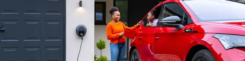 A woman in an orange jumper smiling at a child sitting in a red electric car, with a Pod Point wall charger mounted beside the garage