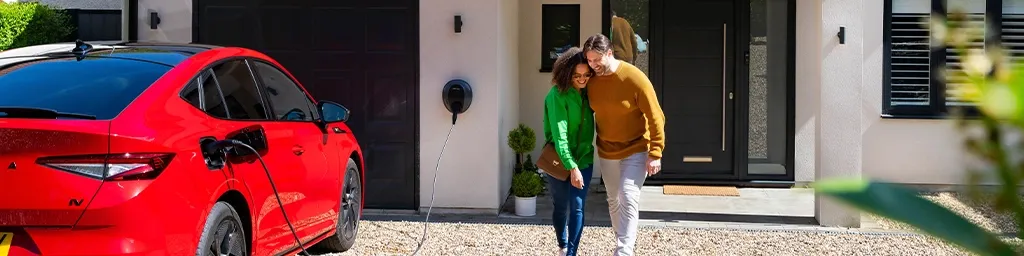 A couple smiling and walking together in front of their home, with a red electric car plugged into a Pod Point wall charger on the driveway