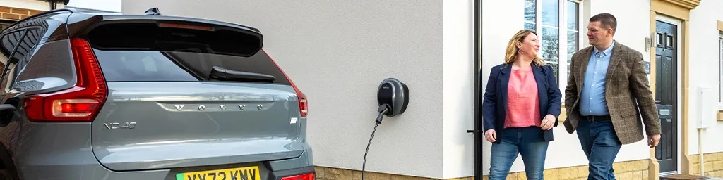 A grey Volvo XC40 electric car charging via a wall-mounted Pod Point home charger, with a couple smiling and walking towards their front door