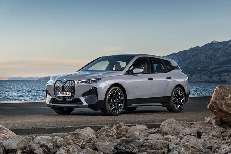 Silver BMW iX electric SUV parked on a road near the coast, with rocky terrain in the foreground and mountains in the background. The sky is clear, and the vehicle is captured in a dynamic, side-on view.