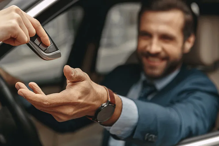 A close-up of a hand passing a car key to a smiling man in a blue suit, who is sitting in the driver's seat of a car. The man extends his hand to receive the key, and the image focuses on their hands with a blurred background.