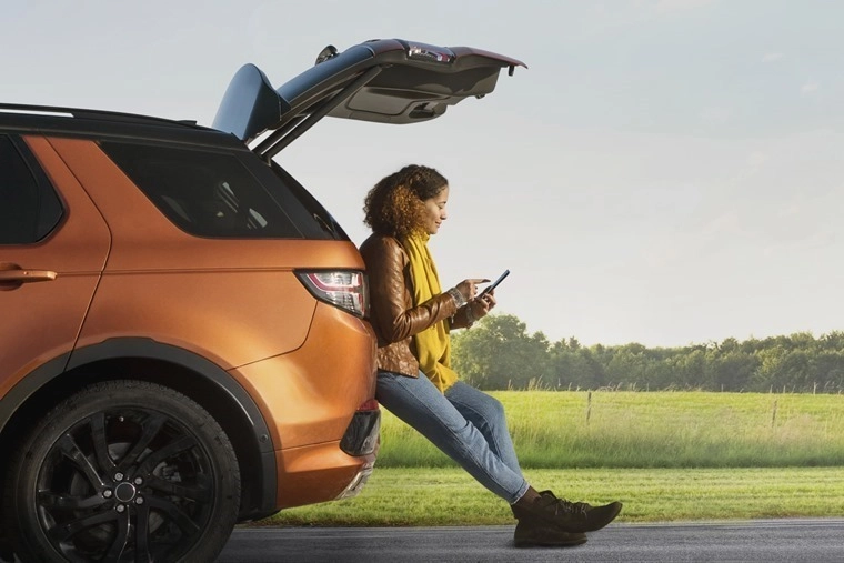 A person seated on the open tailgate of an orange SUV, looking at a mobile device while parked beside a grassy field