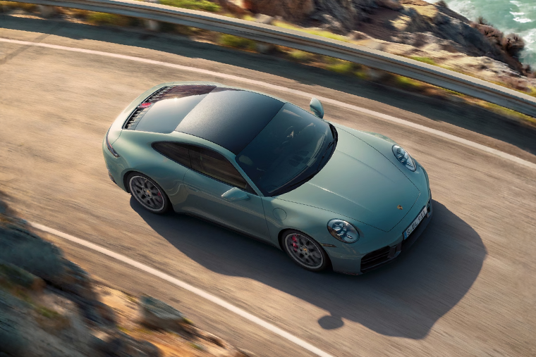 Blue Porsche 911 sports car driving along a winding coastal road viewed from above.