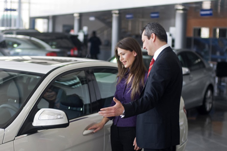 A car salesperson gestures toward a white car for a female customer in a dealership showroom