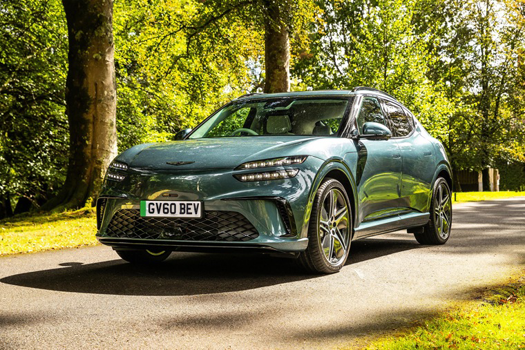 Blue-grey Genesis GV60 electric SUV parked on a paved driveway lined with trees.