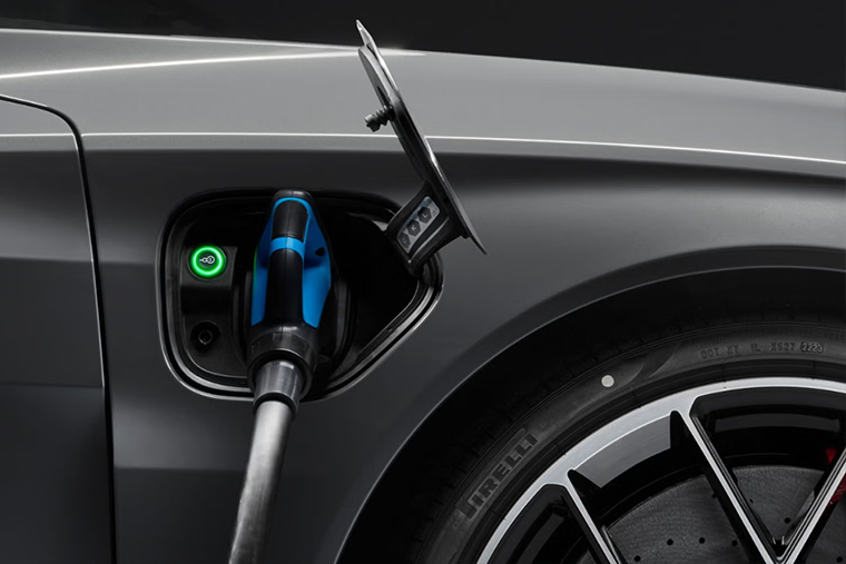 Close-up view of an Audi e-tron charging port with a charging cable connected, showing the illuminated charging indicator and front wheel arch of the vehicle.