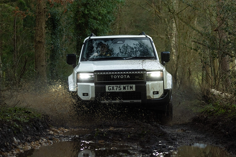 White Toyota 4x4 drives through a muddy woodland trail, headlights glowing as water splashes around its tires; rugged, adventurous mood.