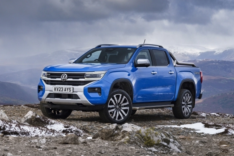 Blue Volkswagen Amarok pickup truck parked on a rocky, snow-dusted mountain ridge, with dramatic clouds and distant peaks behind it.