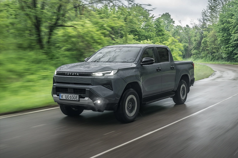 Gray Toyota pickup truck drives along a wet, winding forest road, headlights on, with lush green trees and motion-blurred scenery.
