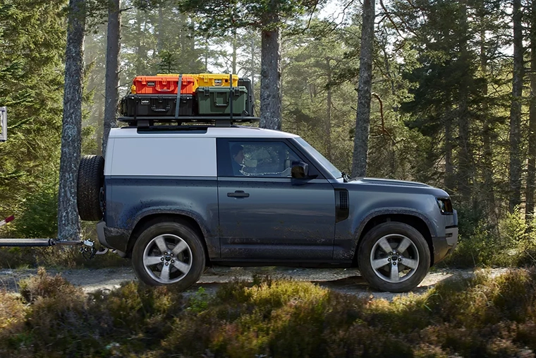 Gray hardtop pick-up with a white roof parked on a forest road, carrying three large cargo containers on its roof rack.