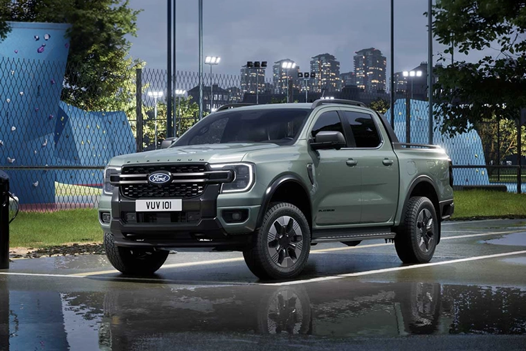 Silver Ford pickup truck parked on a rain-soaked street, reflecting city lights, with a sports field, stadium fencing, and skyline behind it.