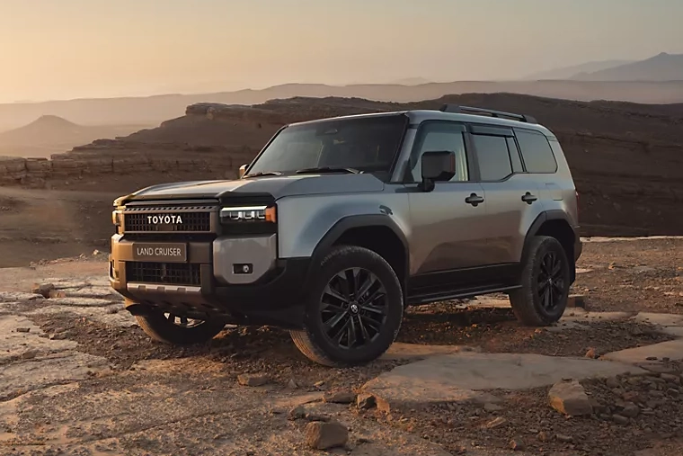 Gray Toyota Land Cruiser pick-up parked on a rugged rocky plateau at sunset, with barren mountains and hazy desert horizons behind it.