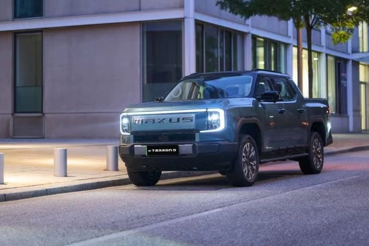 A blue Maxus eTerron 9 pickup truck parked on a city street at dusk, featuring distinctive LED headlights and a bold, modern front grille design.