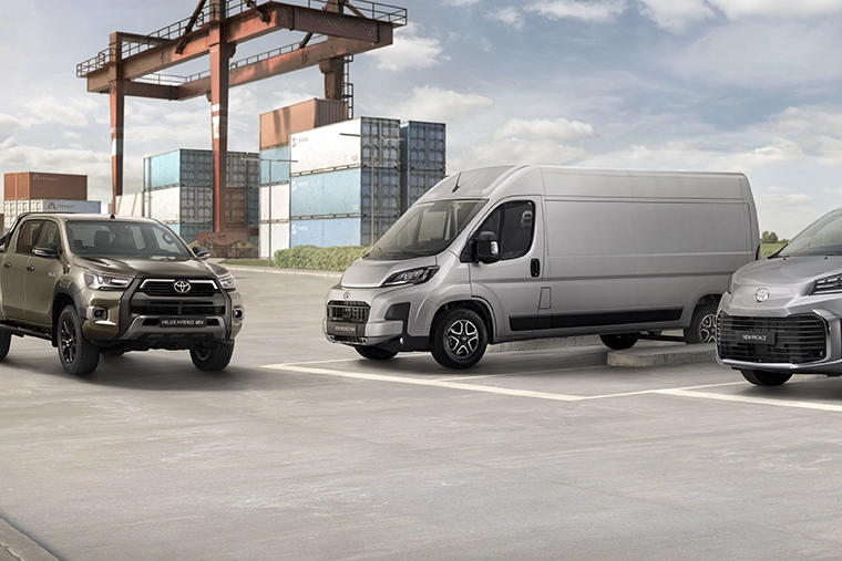 A Toyota pickup, white delivery van, and gray passenger van parked at a shipping container yard beneath a cargo crane.
