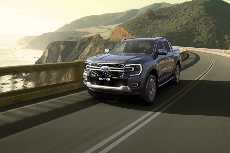 A dark gray Ford Ranger pickup truck drives along a coastal road with a stone railing, overlooking cliffs and the ocean under a soft sunset sky.