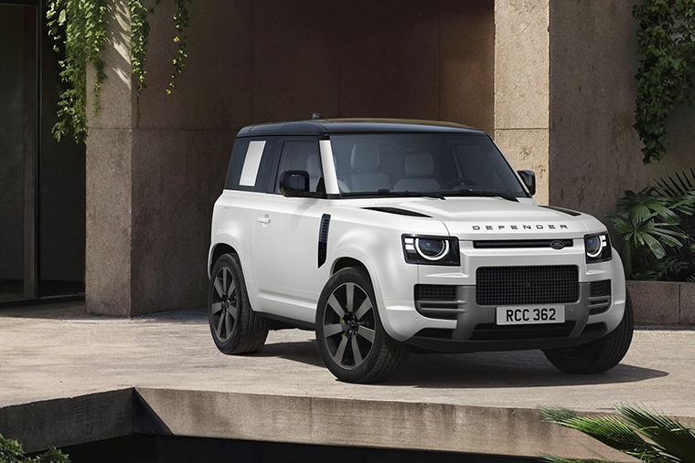 A white Land Rover Defender 90 hardtop with a black roof parked on a modern concrete patio in front of a stone building with hanging green plants.
