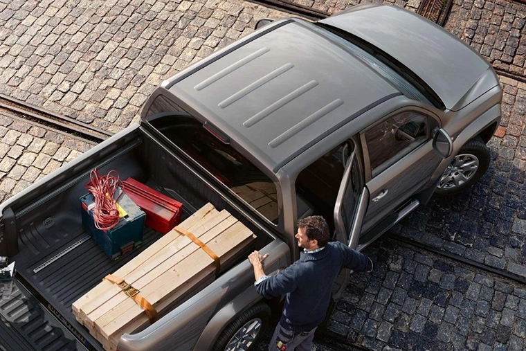 A man stands by the open door of a double-cab pick-up next to a truck loaded with wooden planks, a toolbox, and cables on a cobblestone street.