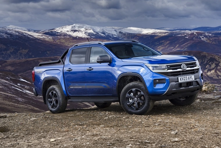 A blue Volkswagen Amarok pickup truck parked on a rocky mountain ridge with snow-capped peaks and a cloudy sky visible in the background landscape.
