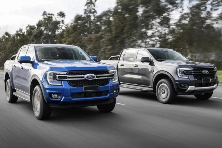 Blue and gray Ford pick-up trucks drive side by side on a road, captured in motion against blurred trees, conveying speed and ruggedness.