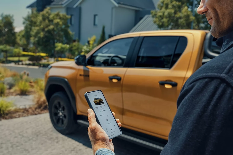 Person holds a smartphone displaying a vehicle app beside an orange pick-up truck in a sunny suburban driveway, conveying connected convenience.