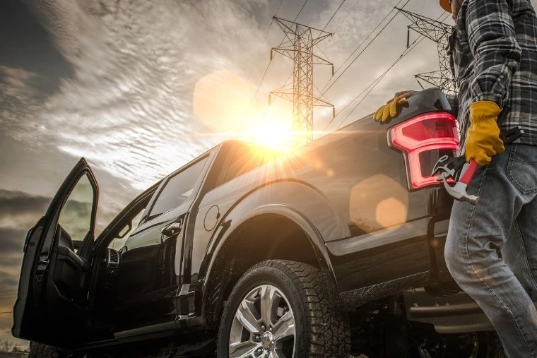 Worker in yellow safety gloves stands beside a black pick-up truck with an open door beneath power lines, bathed in a dramatic sunset.
