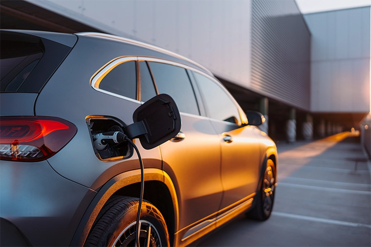 Electric car plugged into a charging station, parked beside a modern building with warm sunset light reflecting off the vehicle’s side.
