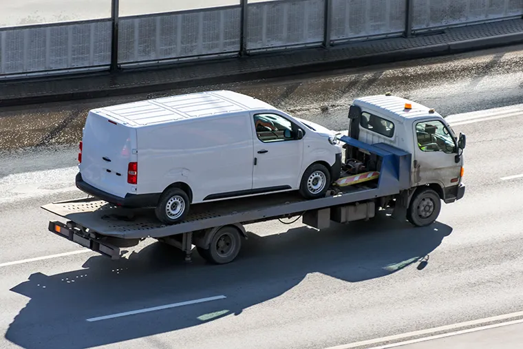 A white van being transported on a flatbed tow truck along a road. Ideal for businesses considering van leasing, showcasing reliable vehicle transport services for leased vans.