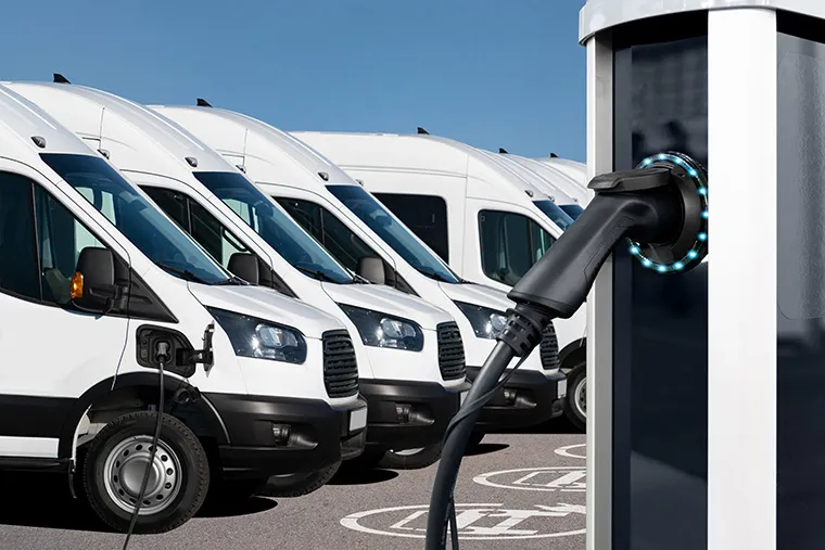 A row of white electric vans being charged at a charging station. The image highlights the convenience and sustainability of electric van leasing, with a close-up of a van plugged into the charger, showcasing a clean and modern fleet for businesses.