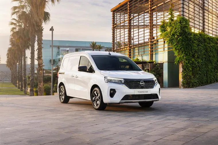A white Nissan Townstar van parked in front of a modern building with palm trees and green landscaping. Ideal for businesses looking for a versatile and stylish van leasing option, combining practicality with sleek design.