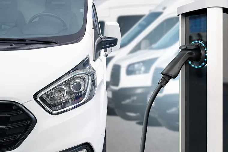 Close-up of a white electric van plugged into a charging station, with a blurred background showcasing additional vans.