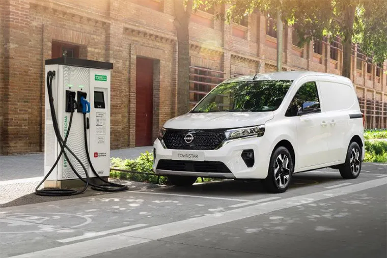A white electric van parked at a public charging station, with the charging cable plugged in. The van is positioned on a city street with modern buildings in the background, emphasizing the convenience and sustainability of electric van leasing for businesses.