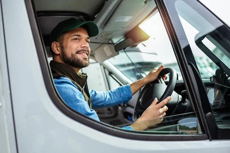 A smiling man wearing a green cap and denim jacket, sitting in the driver's seat of a van. His hands are on the steering wheel, and the vehicle’s interior is visible. Ideal for those interested in van leasing, showcasing a comfortable and practical driving experience.