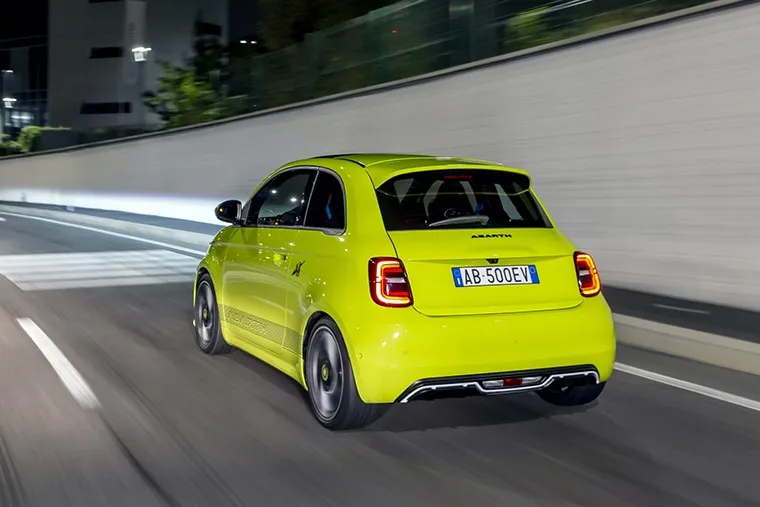 Rear view of a lime green Abarth 500e electric hatchback driving through a city street at night.