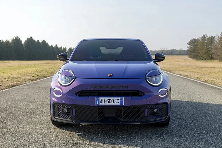 ront view of a vibrant purple Abarth 600e electric car on a quiet country road, showcasing its bold design, LED headlights, and performance-focused stance