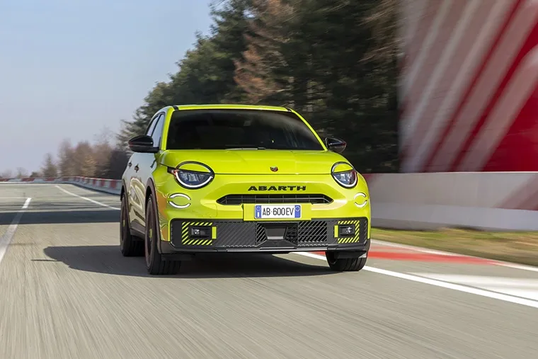 Front view of a vivid green Abarth 600e electric SUV driving on a racetrack, showcasing its bold styling, sporty stance, and aggressive front-end design.