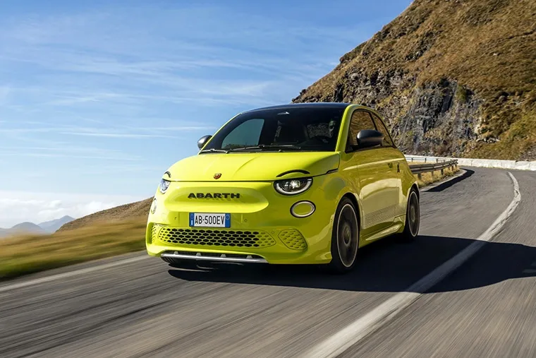 Bright yellow Abarth 500e electric hatchback driving on a winding mountain road under a clear blue sky, highlighting its compact sporty design and agility.
