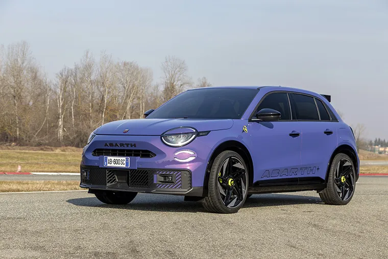 Front three-quarter view of a vibrant purple Abarth 600e compact performance SUV parked on a racetrack with a wooded backdrop.