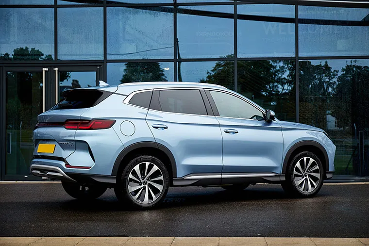 Side profile of a BYD electric SUV in light blue, parked in front of a modern glass building on a wet surface, showcasing its sleek body lines and aerodynamic wheels.