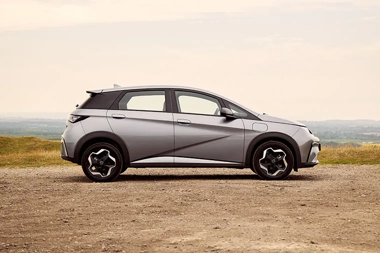 Side view of a silver BYD electric hatchback parked on a gravel surface in an open landscape with rolling hills and a hazy sky in the background.