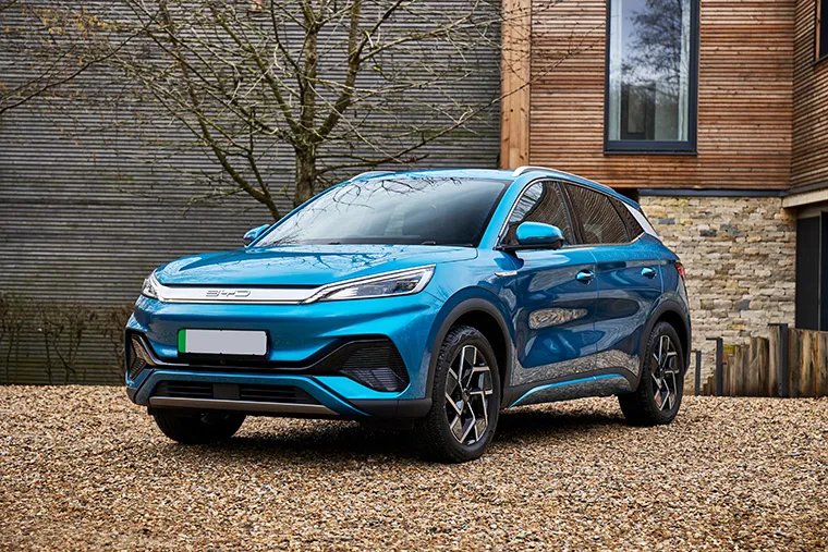 Front three-quarter view of a blue BYD electric SUV parked on a gravel driveway in front of a modern house with stone and wooden exterior walls.
