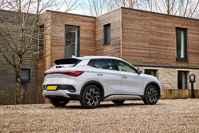 Rear three-quarter view of a white BYD electric SUV parked on a gravel driveway outside a modern home with wooden cladding and a visible EV charger.