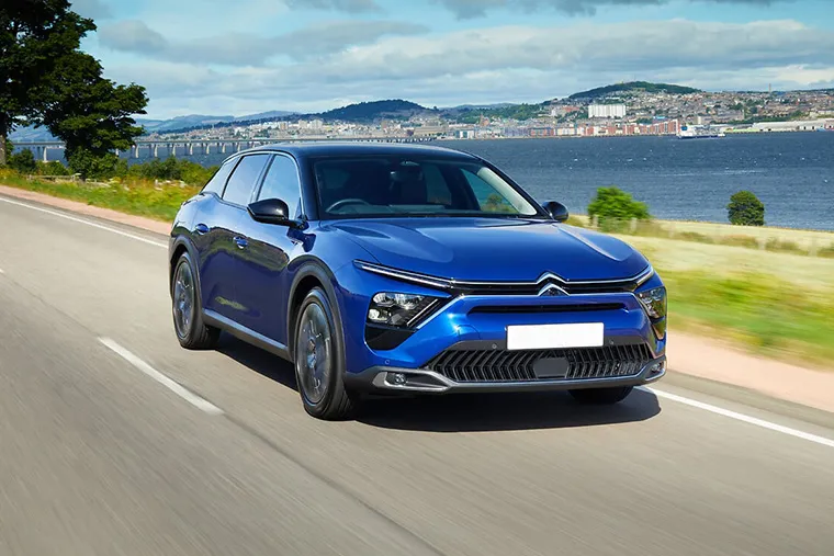 Blue Citroen C5 X SUV driving along coastal road with scenic waterfront and city skyline in background.