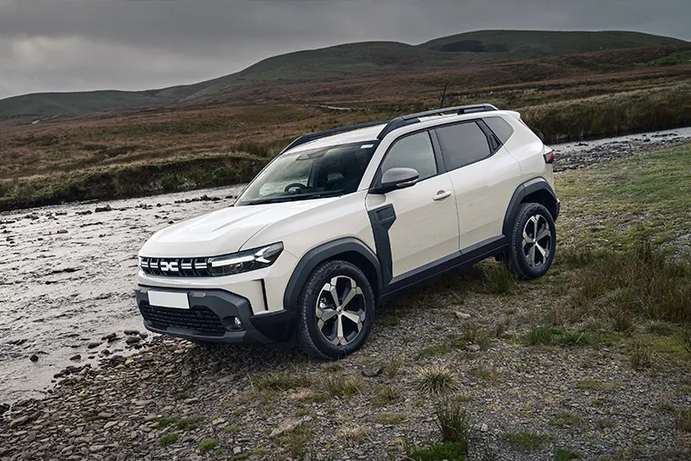 Dacia Duster SUV parked on a rocky riverbank in rugged countryside terrain.