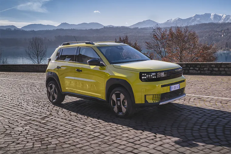 Yellow Fiat Panda electric SUV parked on scenic mountain road with lake and snow-capped peaks in the background.