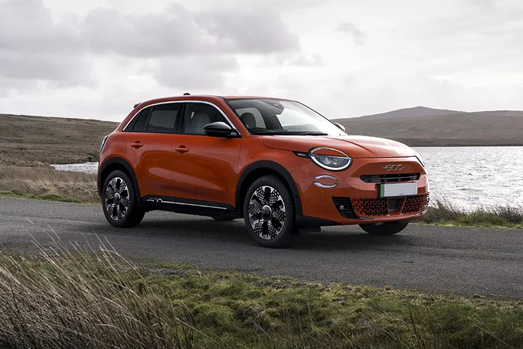 Fiat 600e electric crossover SUV parked by a lake with moody overcast skies and countryside hills.