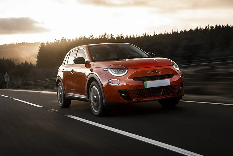 Fiat 600e electric SUV driving on a scenic forest road at sunset.