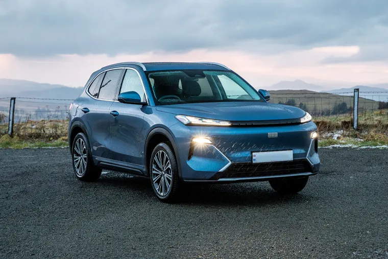 Front three-quarter view of a Geely SUV in metallic blue with headlights on, parked on a gravel road against a moody highland landscape with mountains and cloudy sky
