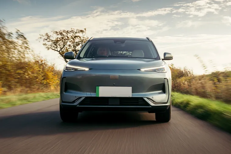 Front-on shot of a Geely electric SUV in grey driving toward the camera on a country road flanked by autumnal hedgerows