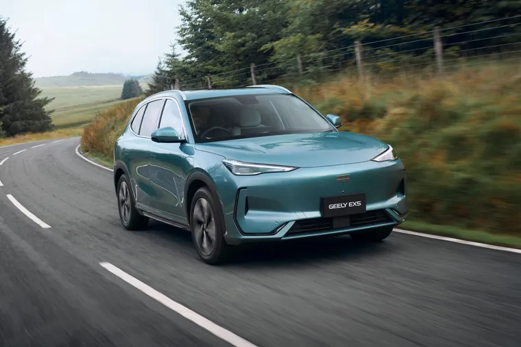 Geely EX5 electric SUV driving on a winding rural road, with motion-blurred hedgerows and trees in the background and green fields visible in the distance