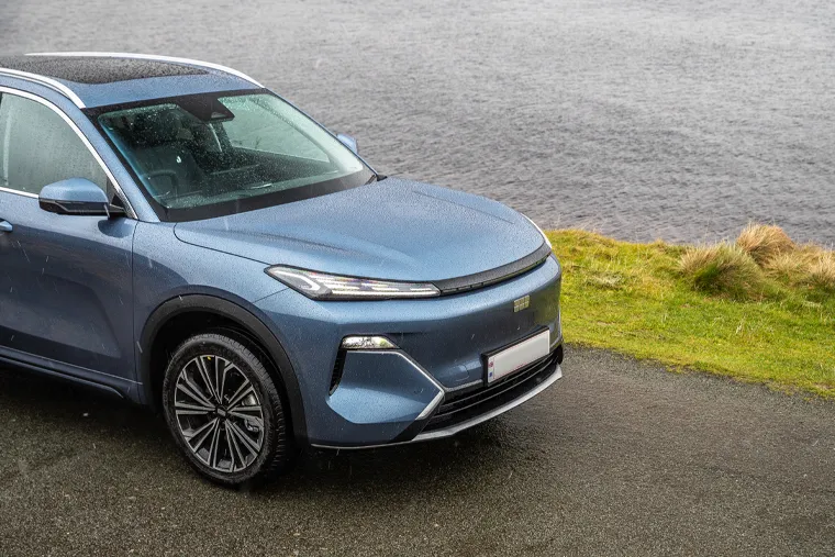 Front three-quarter view of a Geely SUV in metallic blue, parked on wet tarmac beside a grassy bank and body of water